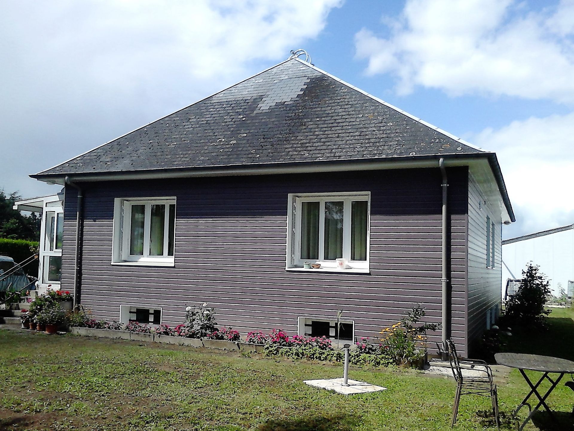 Petite maison grise au toit de bardeaux et parterre de fleurs sous un ciel partiellement nuageux