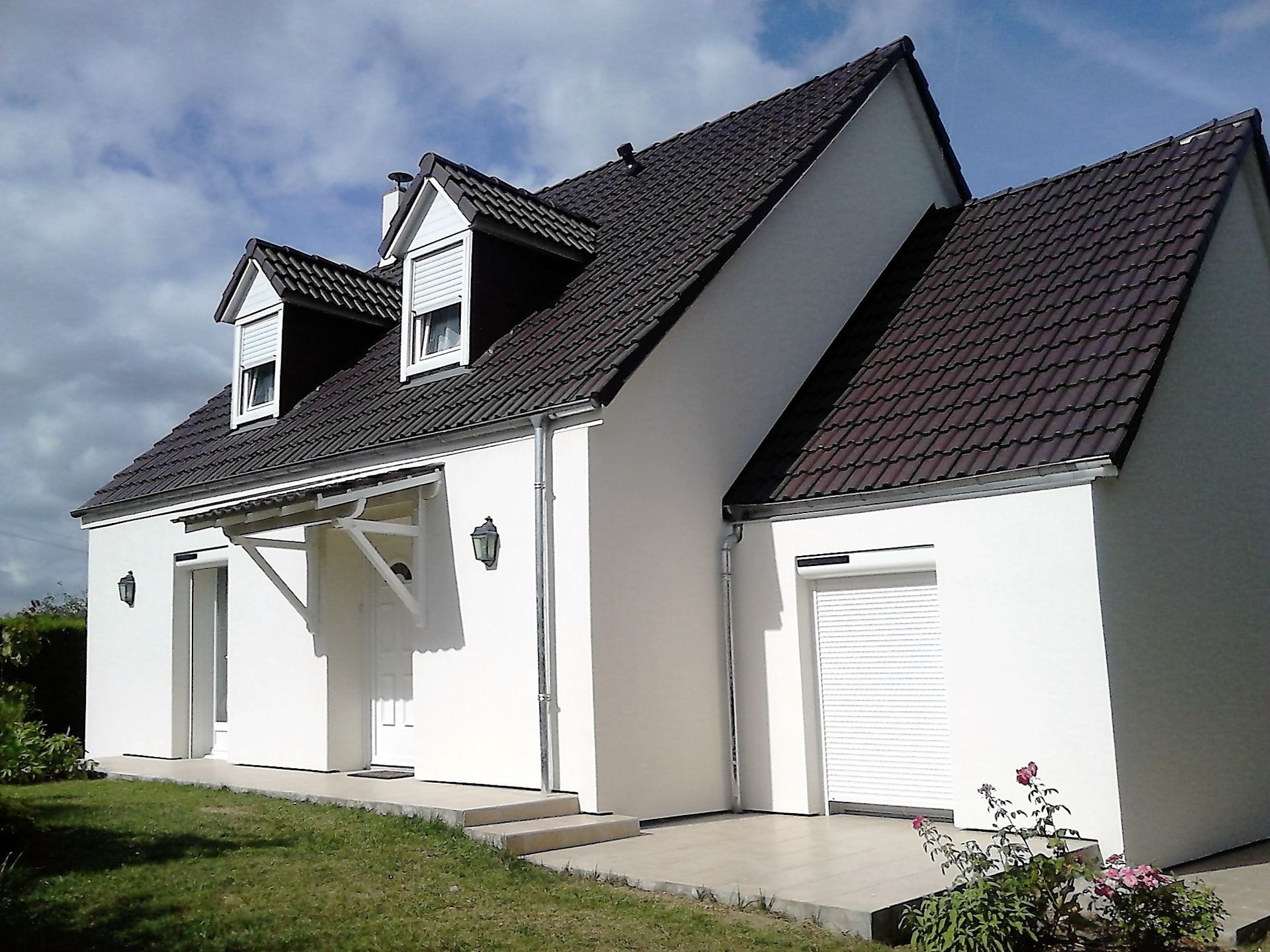 Maison blanche au toit sombre, lucarnes et garage attenant sous un ciel bleu