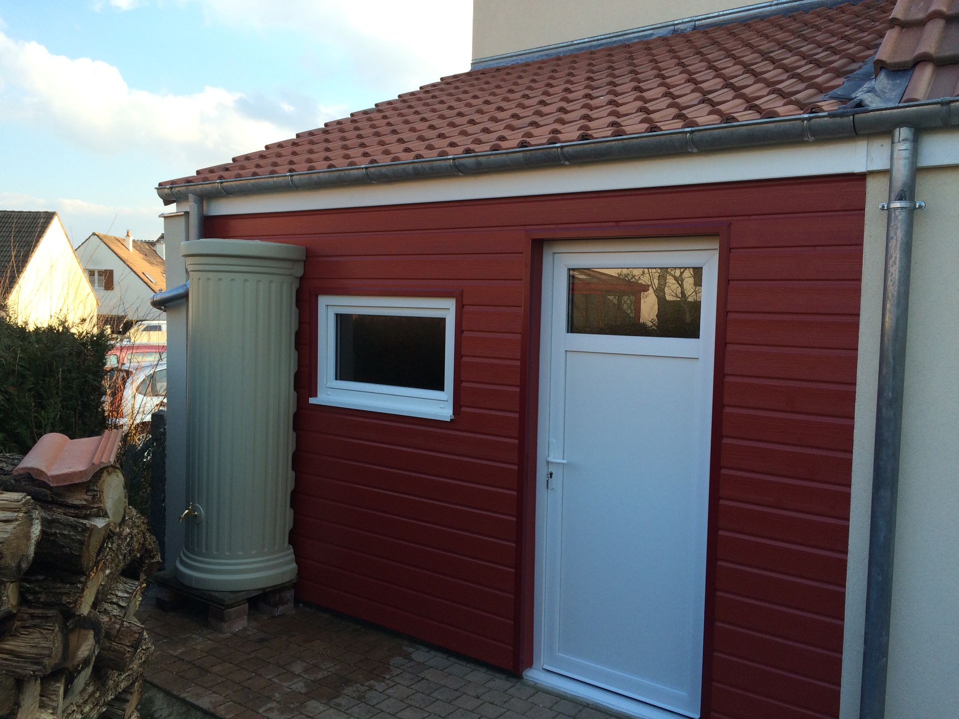 Un hangar en bois rouge avec une porte blanche et une petite fenêtre, à côté d'une grande cuve ronde grise sous un toit de tuiles.