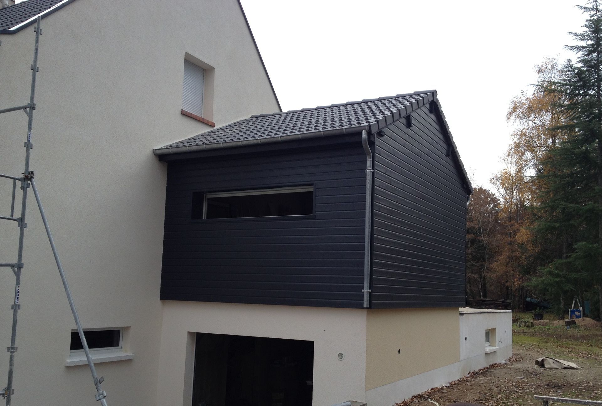 Maison moderne à deux étages avec un bardage foncé et un toit en pente, située à côté d'un bâtiment beige, près d'arbres.