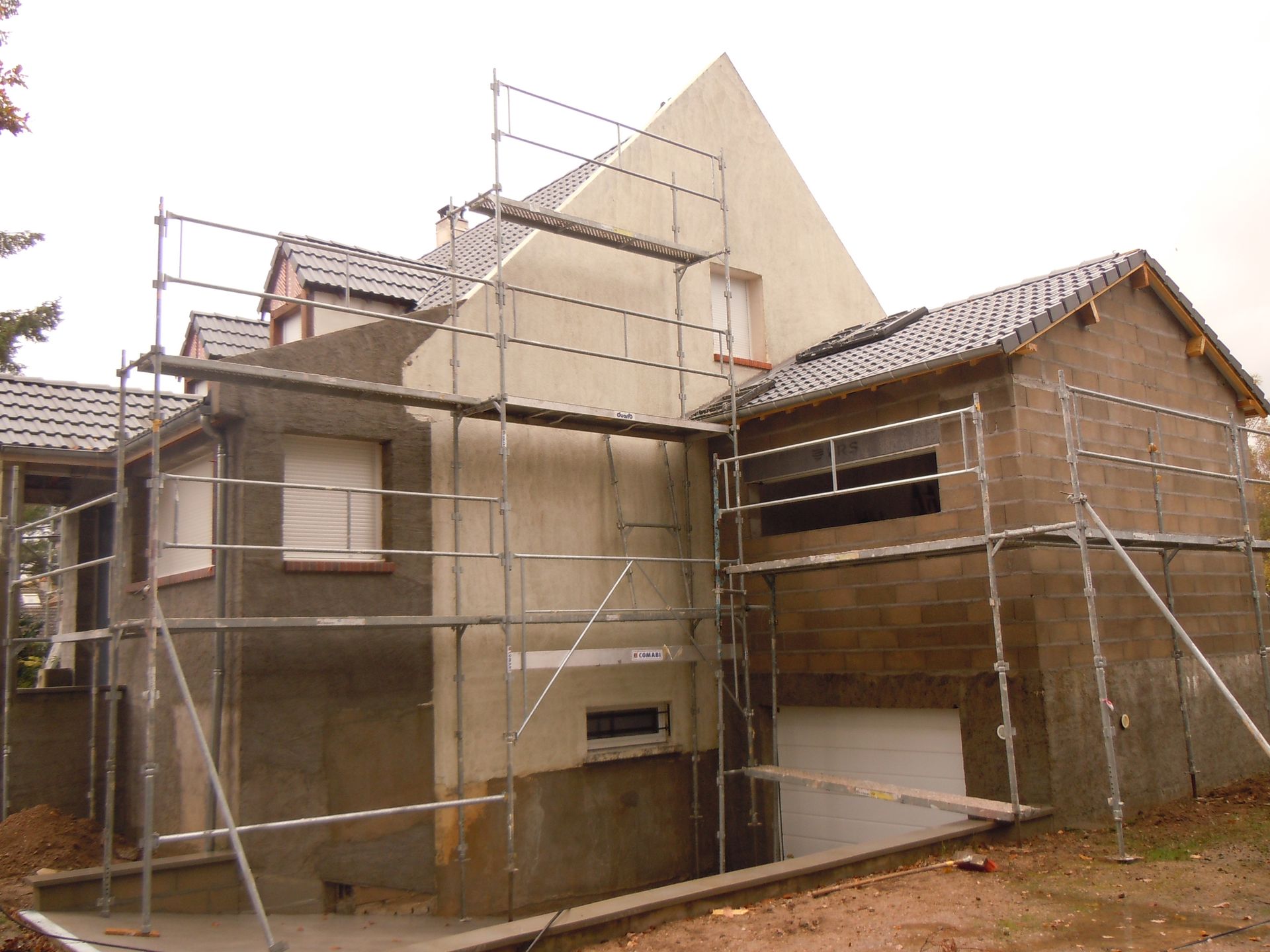 Des échafaudages entourent une maison en rénovation aux murs beiges et au toit de tuiles.