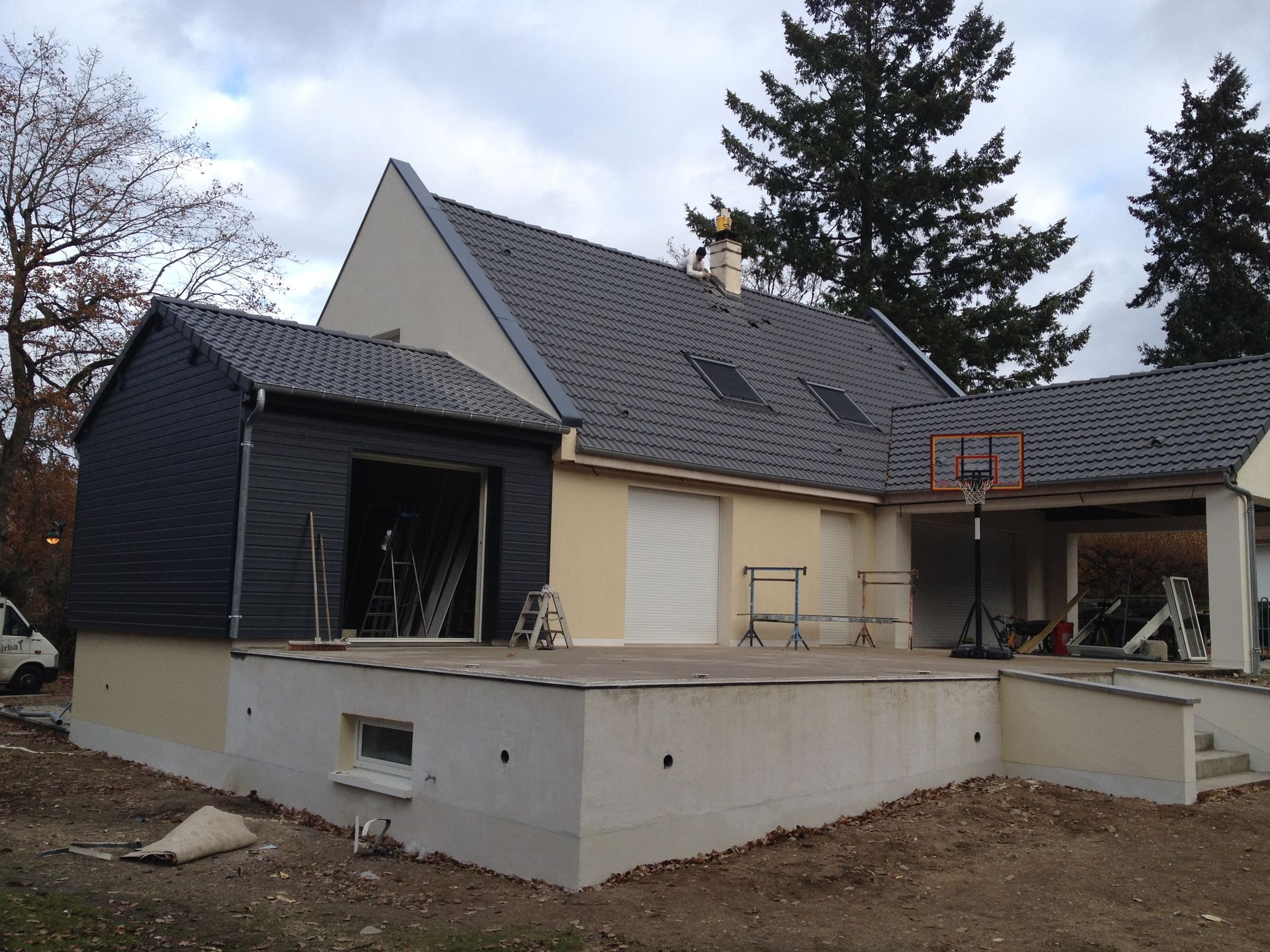 Maison en construction avec murs en stuc beige, toit foncé et patio inachevé dans une cour.
