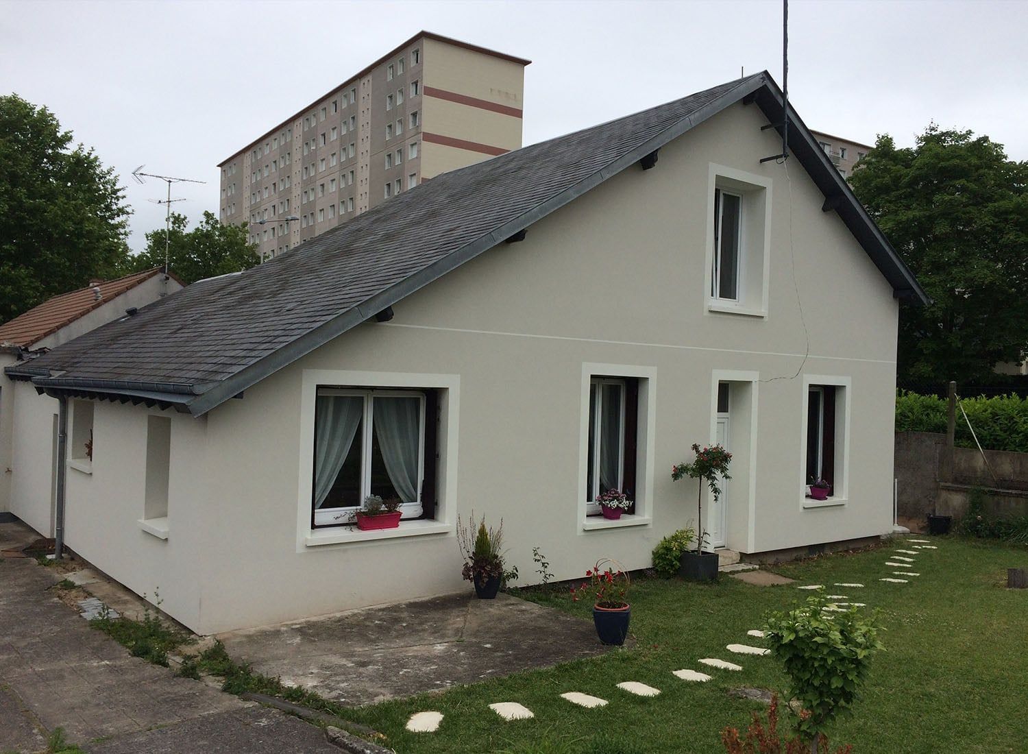 Petite maison couleur crème avec un toit gris foncé, des fenêtres à encadrement blanc et un petit jardin devant.