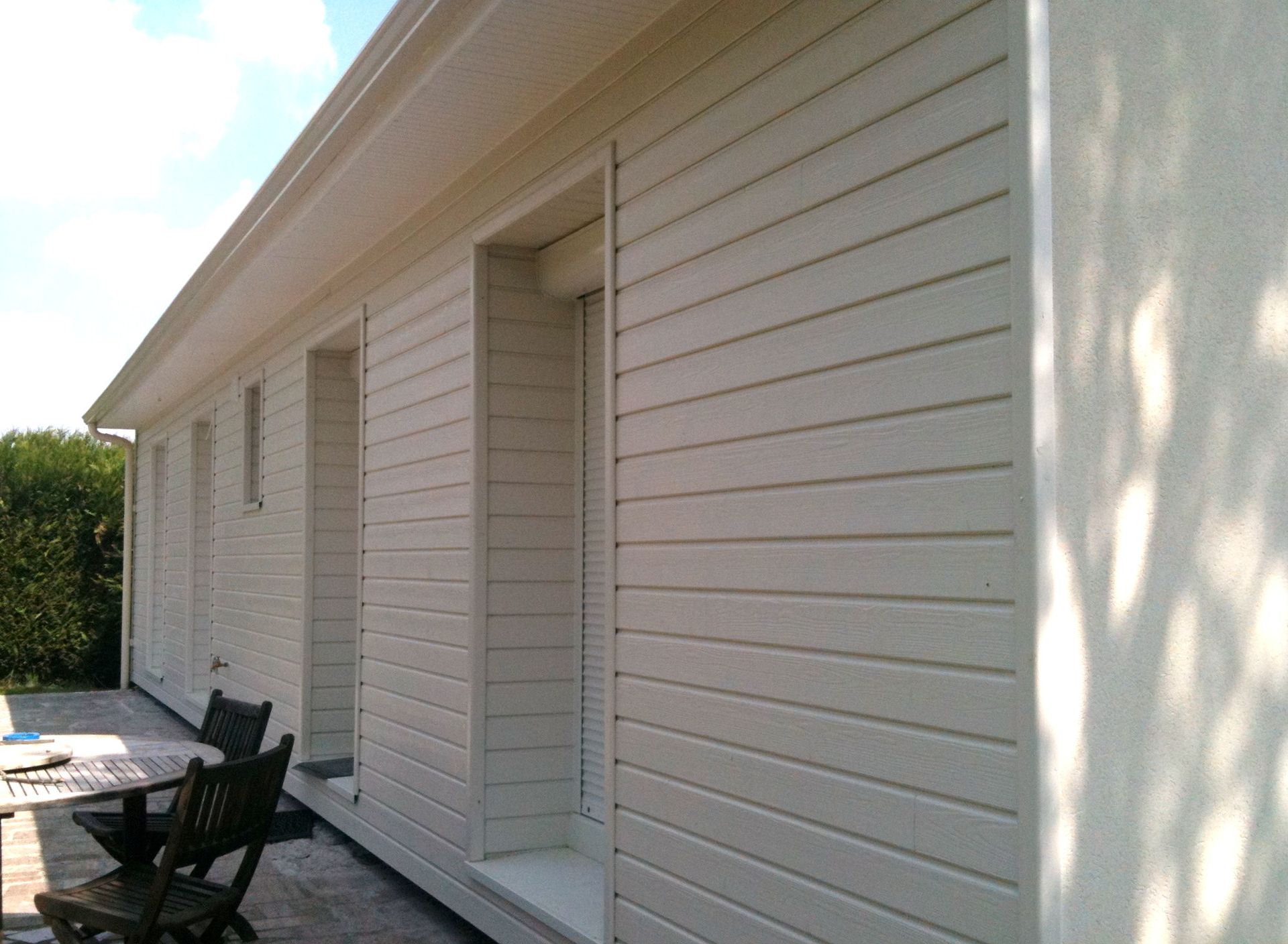 Maison à façade blanche avec terrasse en bois et chaises d'extérieur le long d'une allée étroite.