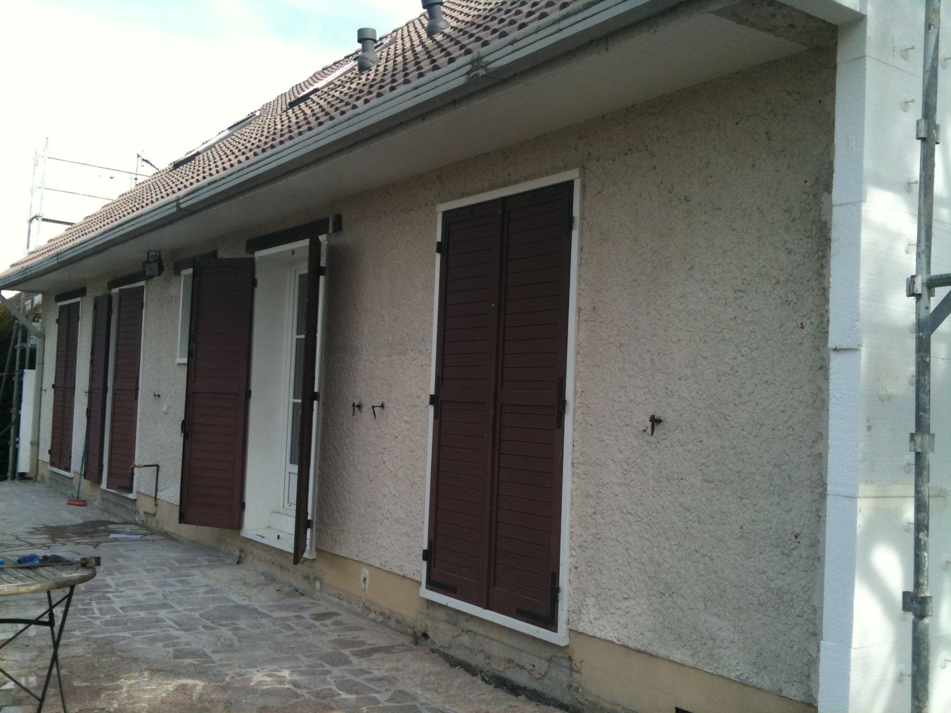 Extérieur d'une maison aux murs en stuc beige, aux volets bruns et au toit de tuiles, le long d'une allée étroite.