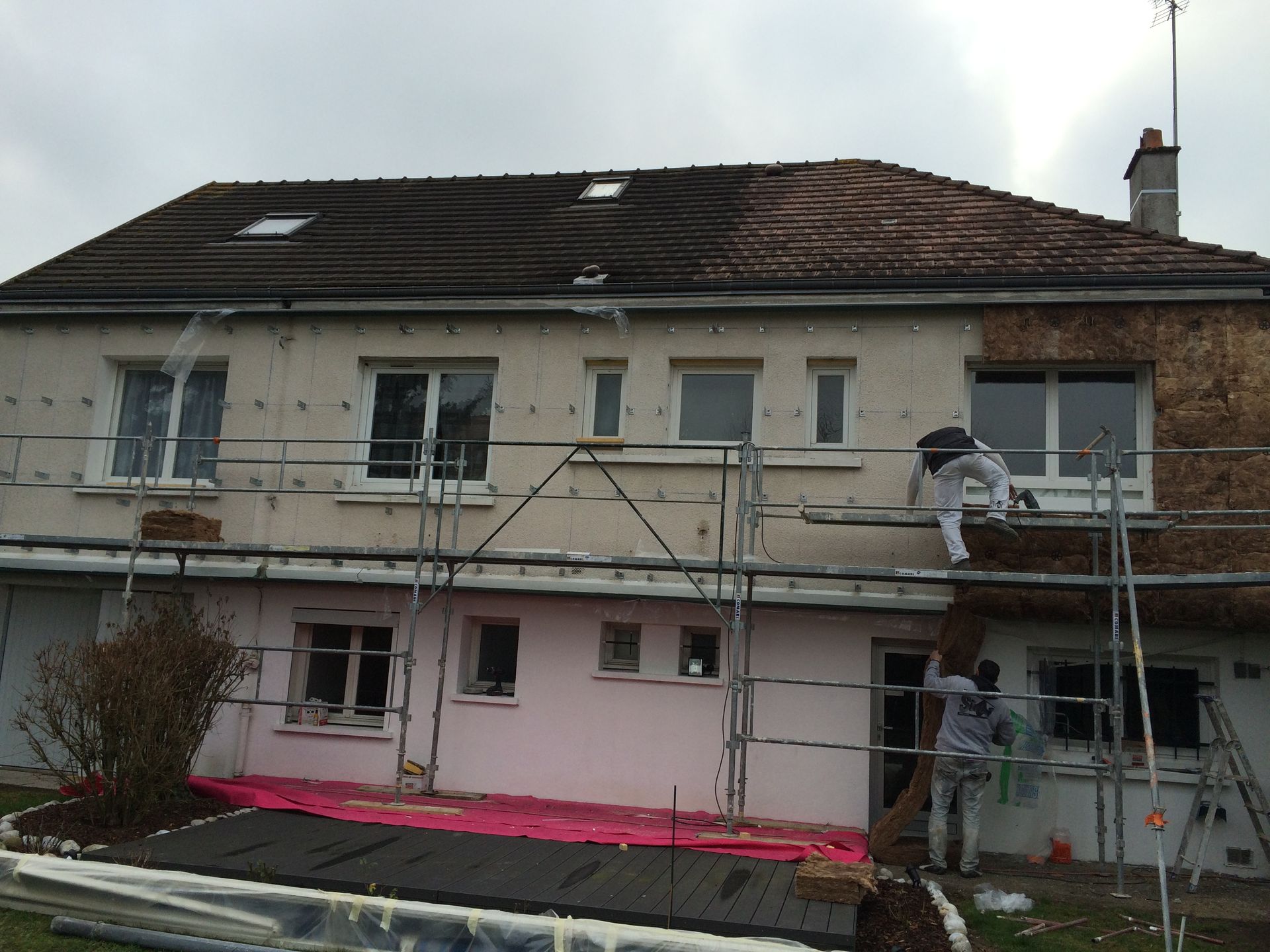 Deux ouvriers sur un échafaudage réparent la façade d'une maison à deux étages dont le toit est endommagé.