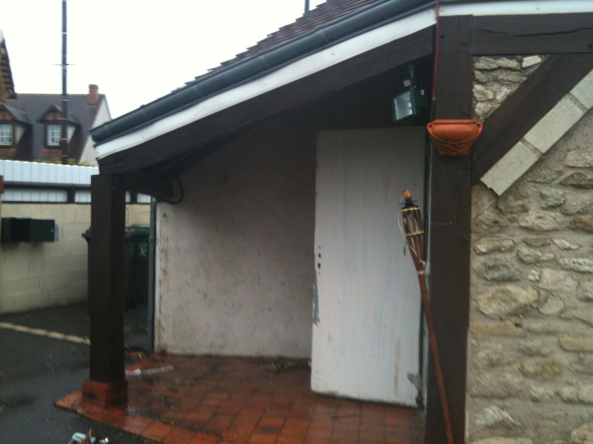 Petit abri de jardin avec une porte blanche et un toit foncé, à côté d'une terrasse en briques humides.