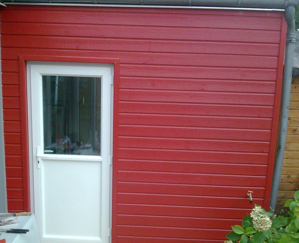 Mur extérieur rouge avec une porte et une fenêtre blanches, jouxtant un petit jardin