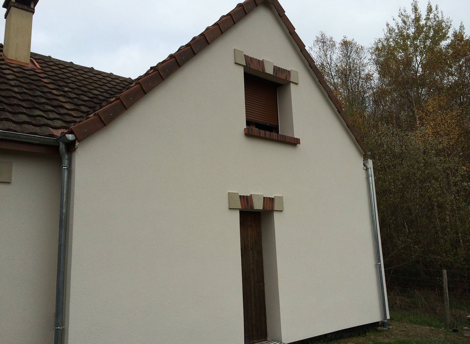 Petite maison blanche au toit pentu et à la fenêtre du haut condamnée, entourée d'arbres.
