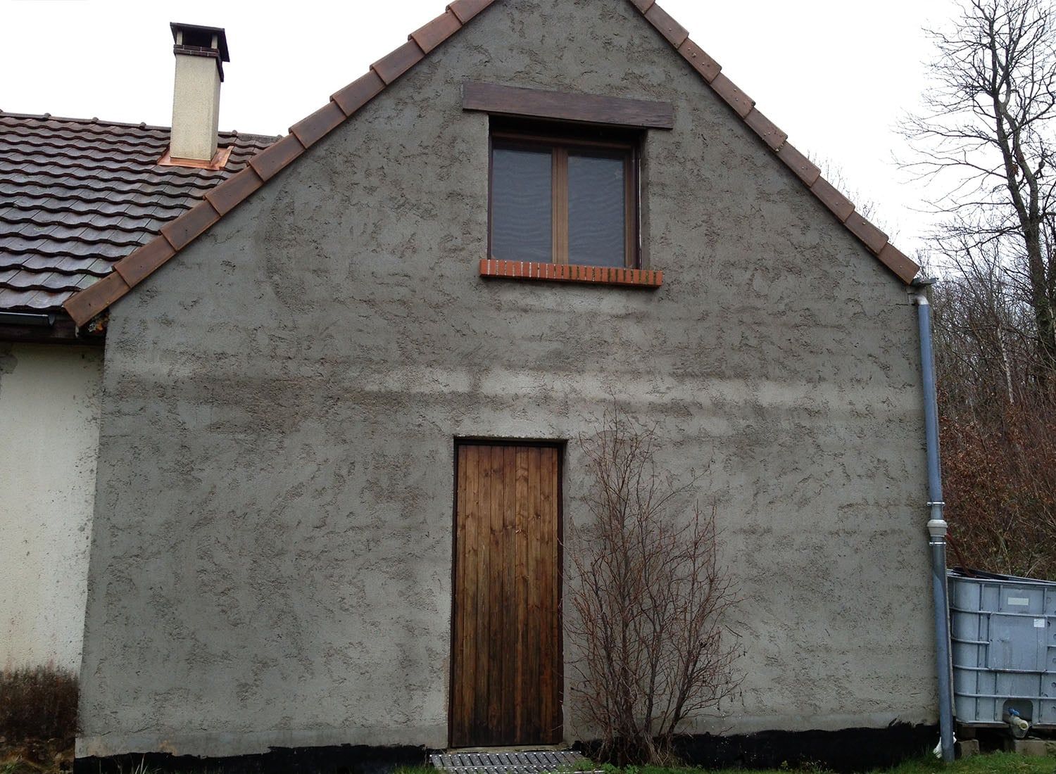 Maison en stuc gris avec porte marron, fenêtre à l'étage et toit de tuiles sous un ciel couvert