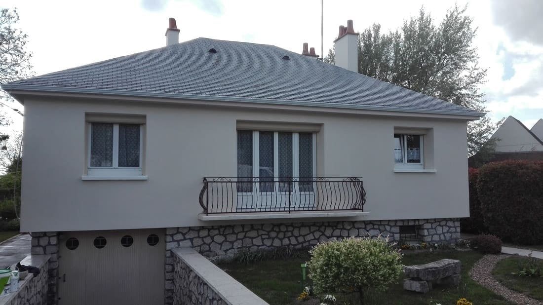 Petite maison de plain-pied avec toit gris, murs blancs, balcon et fondations en pierre derrière un chemin de jardin.