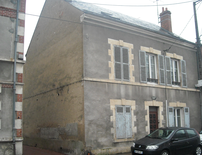Vieille maison grise dans une rue étroite, avec des fenêtres à volets et une voiture noire garée.