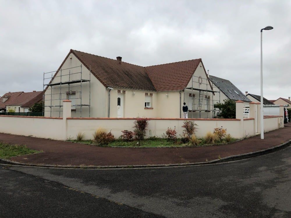Maison blanche au toit de tuiles rouges, dissimulée derrière un muret, à l'angle d'une rue de banlieue balayée par les nuages.