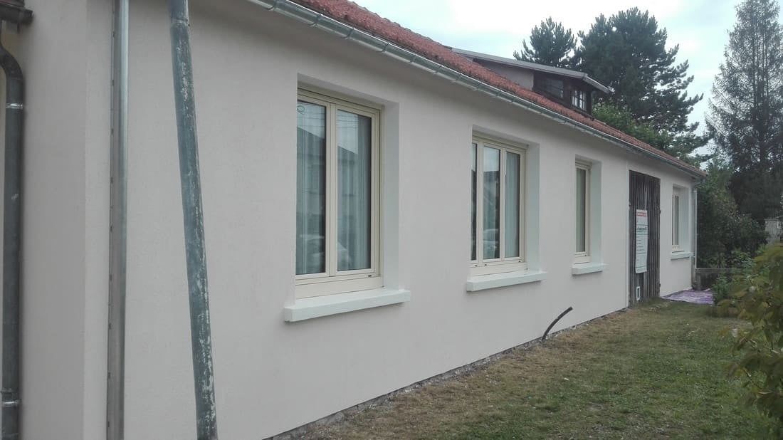 Extérieur d'une maison blanche avec trois fenêtres et un toit en pente, vue de côté dans une cour gazonnée.