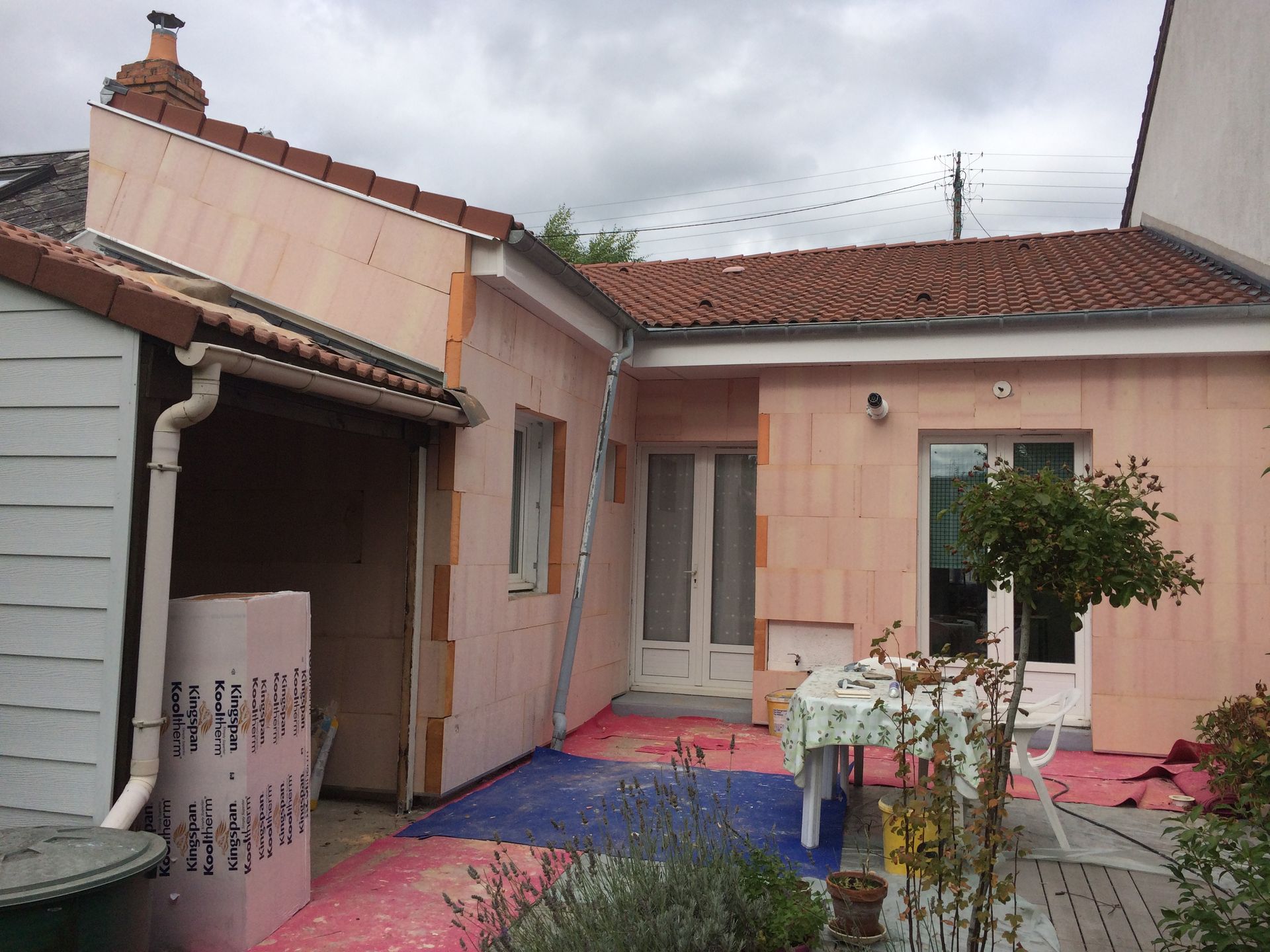 Terrasse arrière avec murs roses, toit de tuiles, tapis colorés et un petit arbre près de la porte.