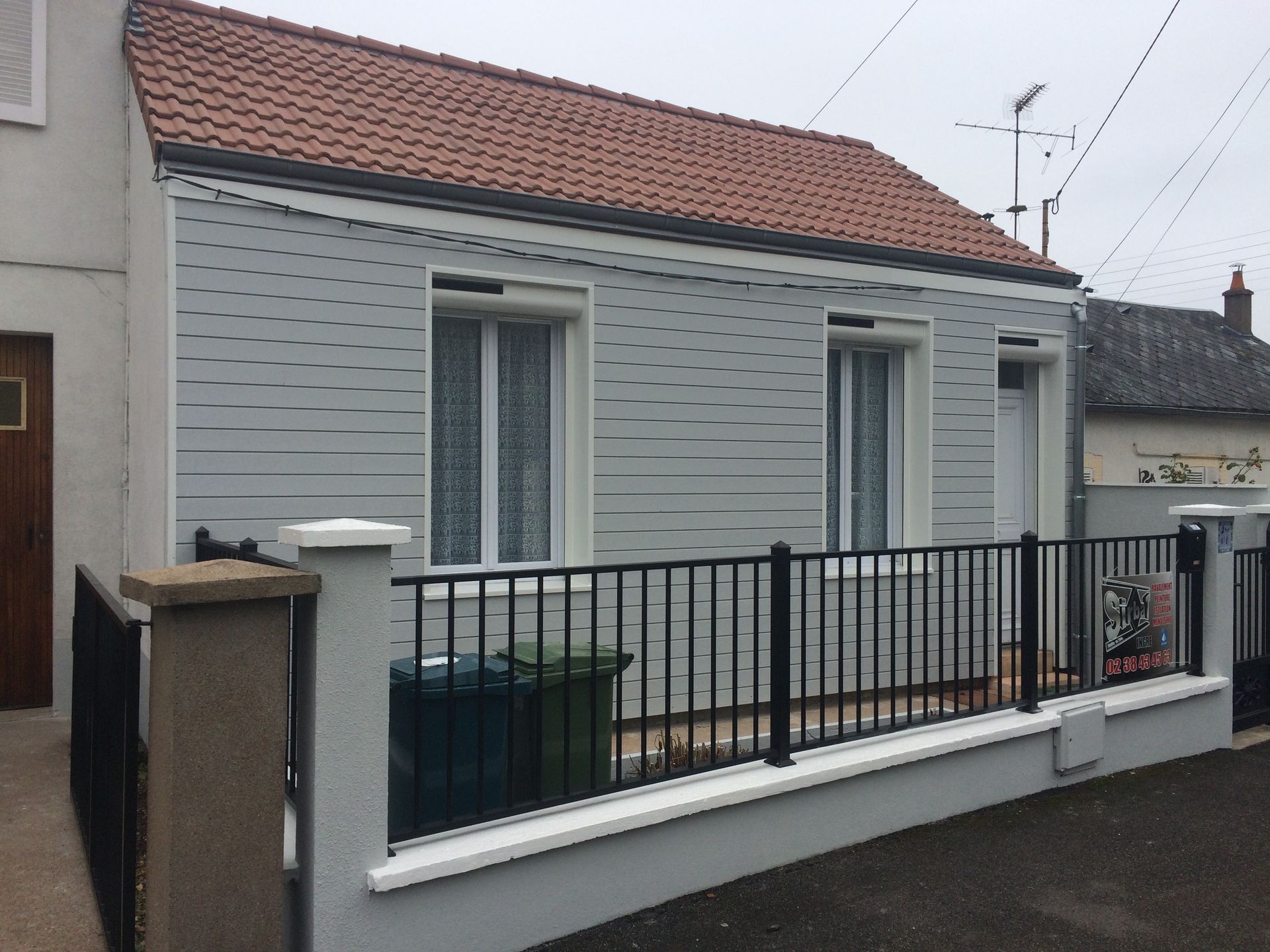 Petite maison grise au toit de tuiles rouges et à la clôture noire, dans une rue résidentielle.