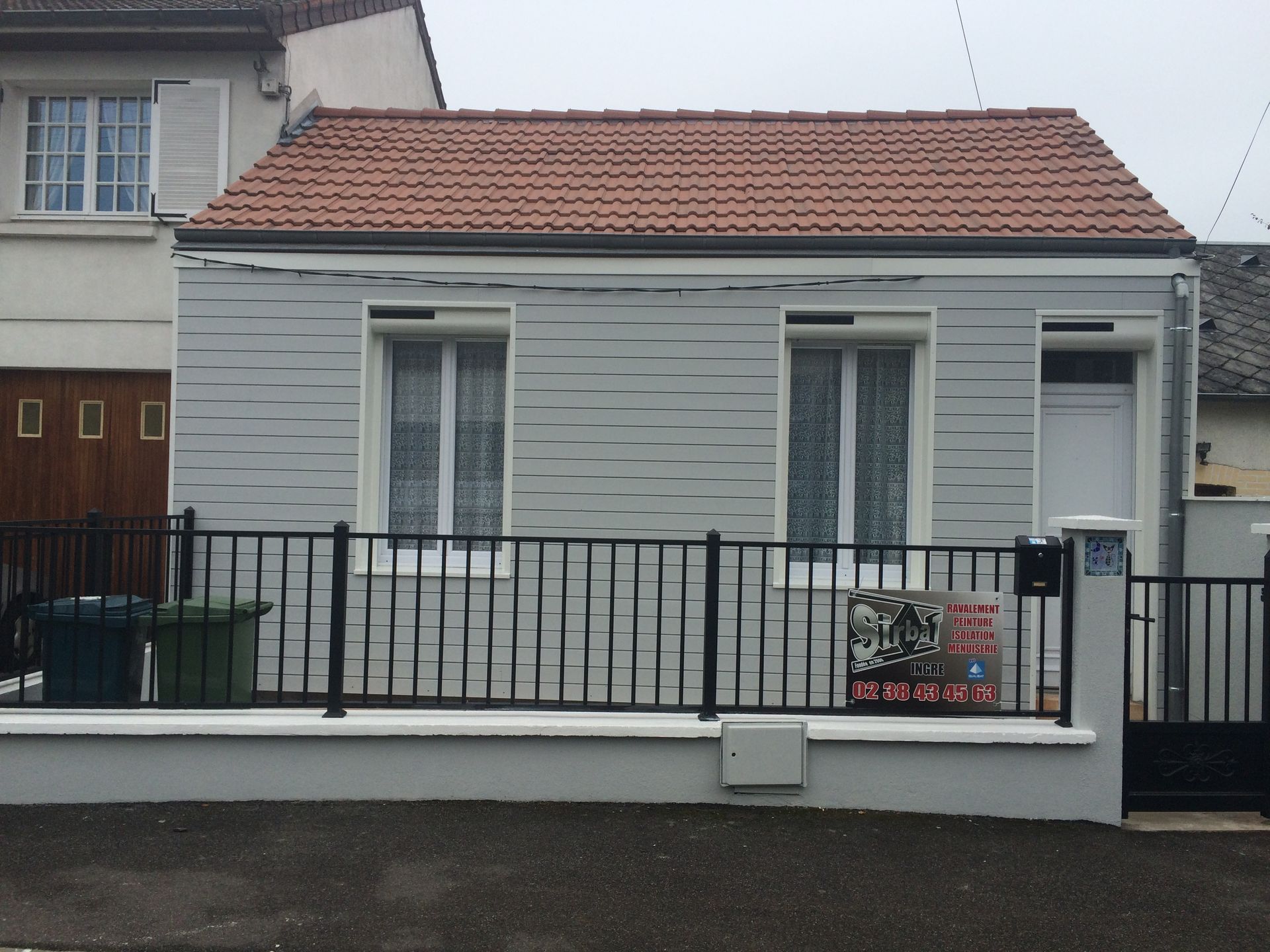 Petite maison blanche avec un toit de tuiles rouges et une clôture en métal noir devant.