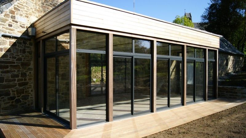 Extension moderne en verre adossée à une maison en pierre, avec toit en pente et terrasse en bois.