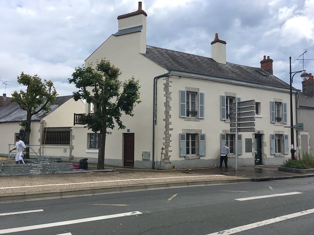 Bâtiment blanc de deux étages aux volets gris, en bordure de route, avec quelques piétons à l'extérieur.