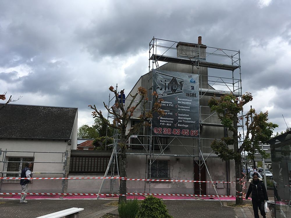 Échafaudages autour d'un bâtiment en rénovation, avec des ouvriers, des échelles et du ruban de signalisation rose devant.