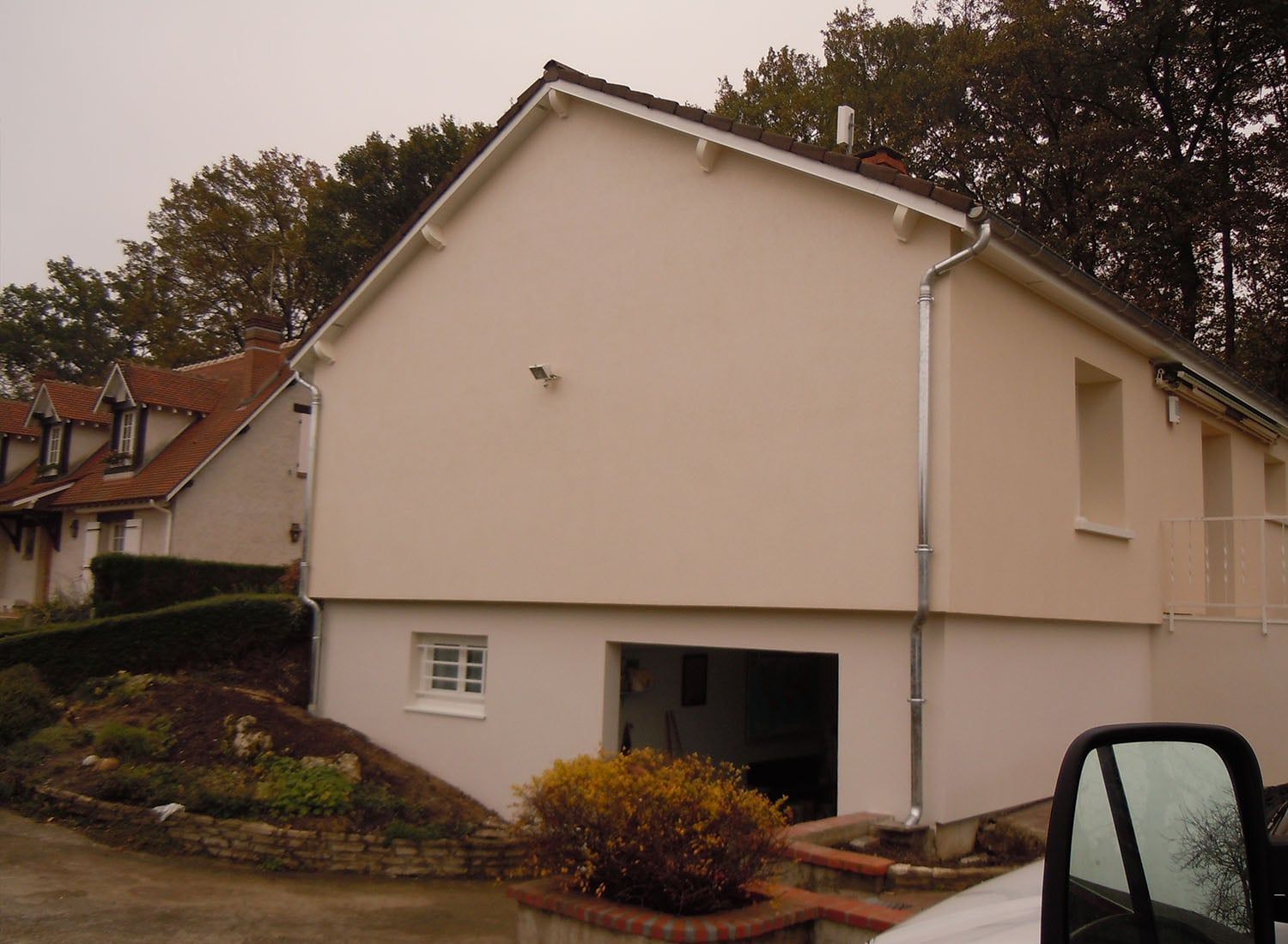 Maison de banlieue aux murs beiges et au toit en pente, vue depuis une allée bordée d'arbres.