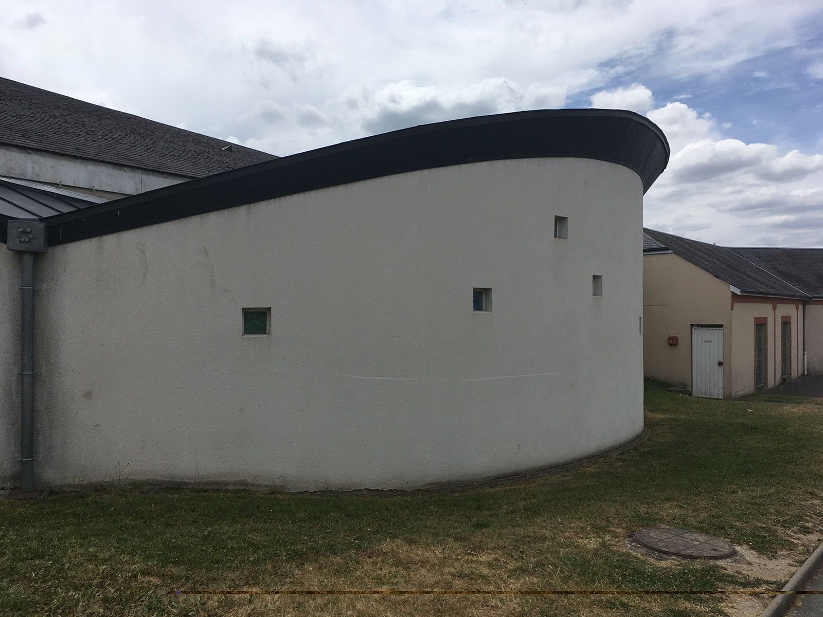 Bâtiment blanc incurvé avec un toit sombre sur un terrain herbeux sous un ciel partiellement nuageux