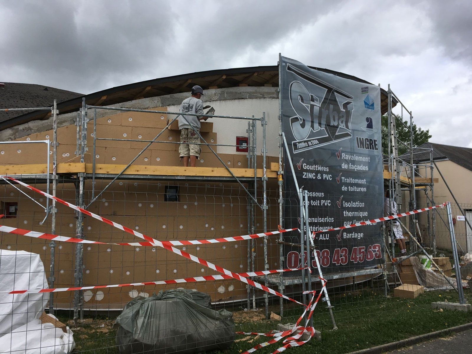 Chantier de construction avec un ouvrier sur un échafaudage à côté d'un bâtiment recouvert de filets de sécurité et de ruban adhésif rouge et blanc