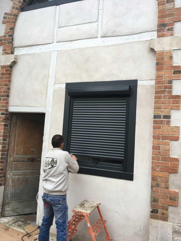 Un ouvrier installe un volet roulant noir sur la façade d'un bâtiment, une petite échelle à proximité.