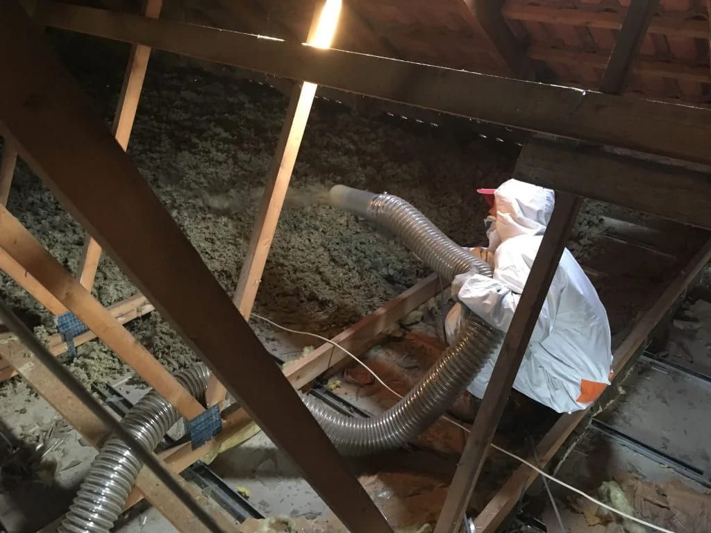 Un ouvrier en combinaison de protection blanche inspecte l'isolation des combles et les conduits flexibles sous les poutres de toiture apparentes.