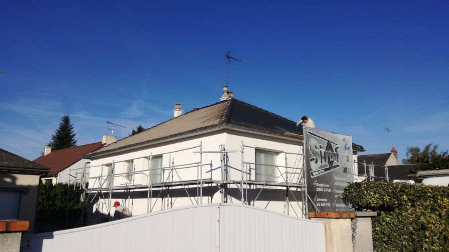 Maison blanche avec échafaudages et travaux de toiture sous un ciel bleu dégagé, visible derrière une clôture.
