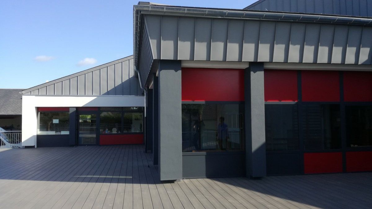 Entrée d'un immeuble moderne avec des panneaux gris, des touches de rouge et une cour pavée sous un ciel bleu azur.