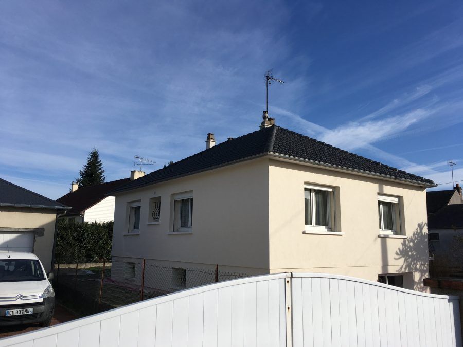 Maison d'habitation au toit sombre et aux murs blancs, vue depuis une cour sous un ciel bleu.
