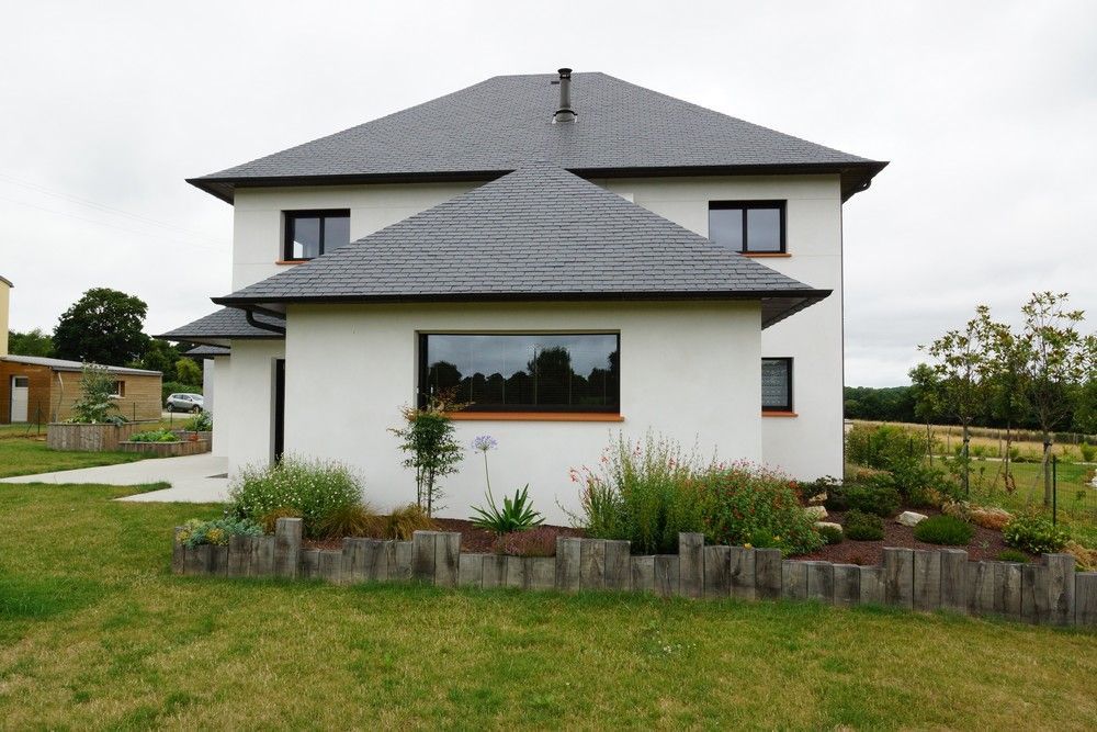 Maison moderne à deux étages avec murs blancs, toit foncé et jardin à l'avant sous un ciel couvert.
