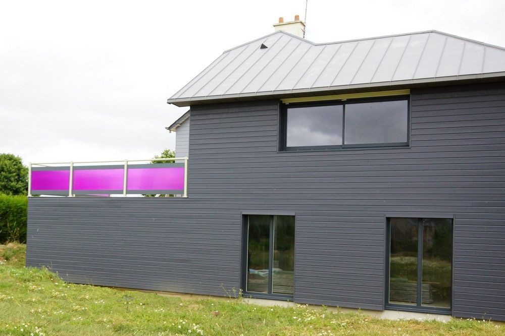 Maison grise moderne avec toit en métal, fenêtres sombres et panneaux de balcon rose vif dans une cour gazonnée