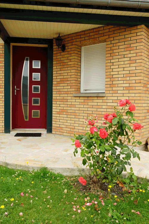Entrée d'une maison en briques avec porte rouge, volets blancs et rosier dans le jardin avant