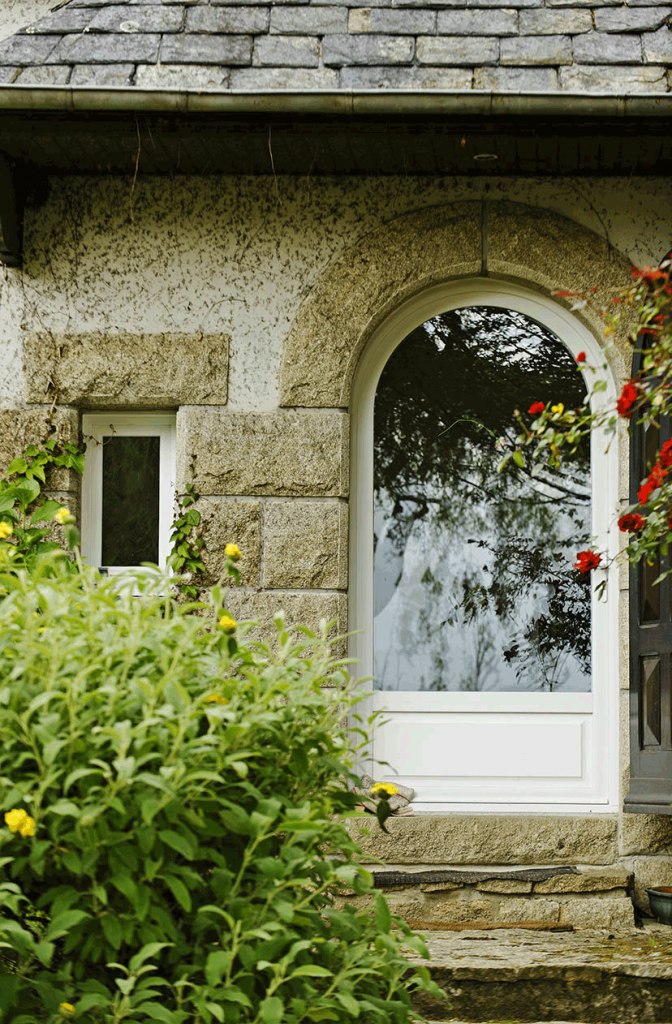 Entrée d'une maison en pierre avec une porte blanche cintrée, une petite fenêtre et de la végétation luxuriante devant.