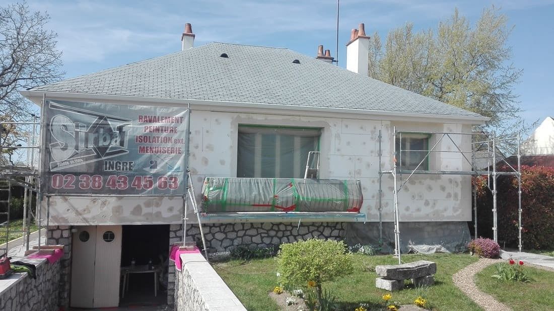 Petite maison blanche au toit gris incurvé, ornée de bannières en façade et d'une entrée au sous-sol, dans un jardin arboré.