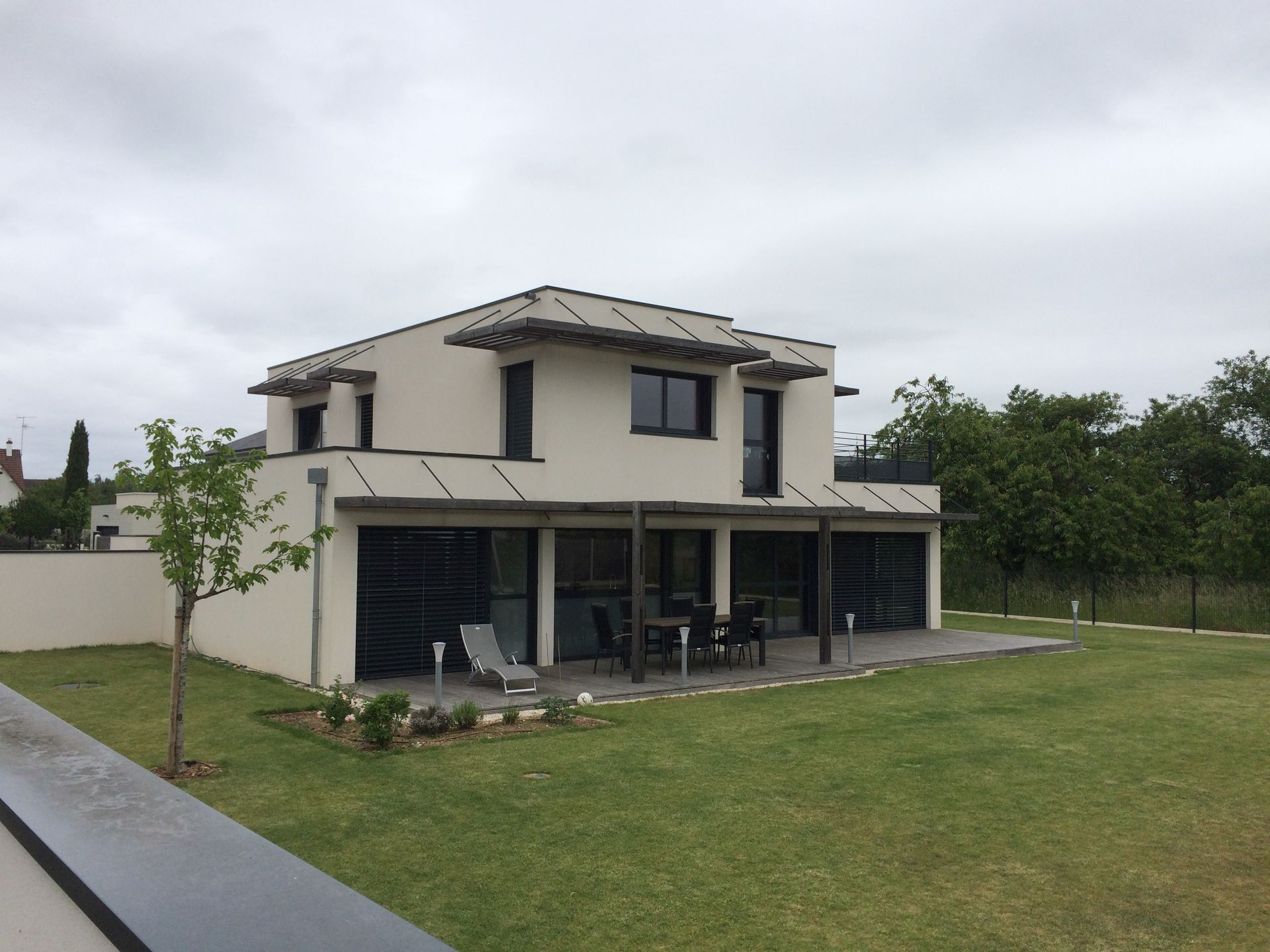 Maison moderne à deux étages avec murs couleur crème, toit foncé, mobilier de patio et grand jardin gazonné.