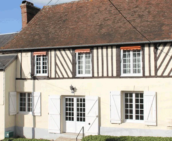 Maisonnette blanche à colombages noirs et blancs, toit rouge et fenêtres à volets baignées de soleil