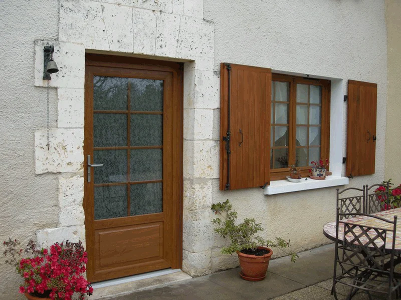 Façade de maison en pierre avec porte en bois, fenêtre à volets, plante en pot, table et chaises de patio