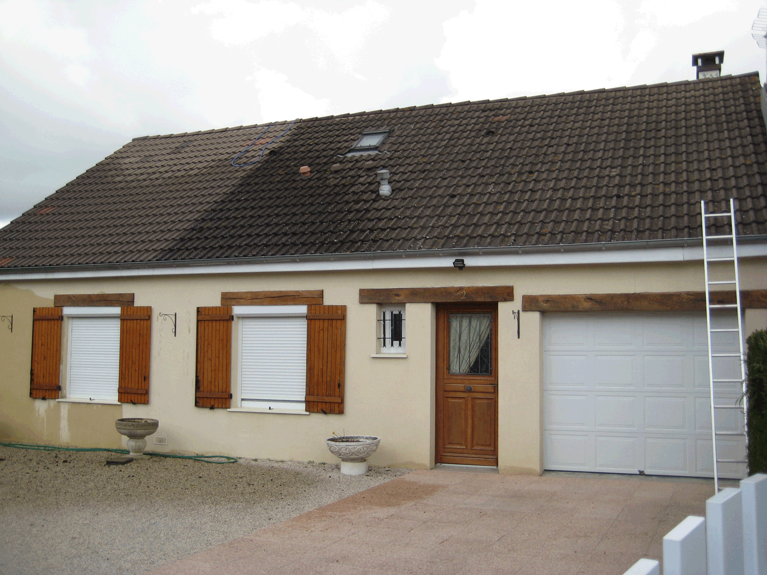 Maison de plain-pied avec des volets marron, une porte de garage blanche et une échelle appuyée contre le toit.