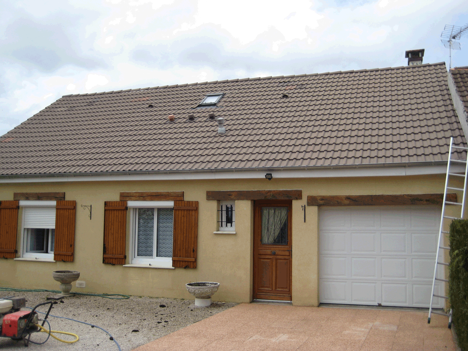 Maison de plain-pied avec volets marron, toit de tuiles, garage et échelle sur le côté droit.