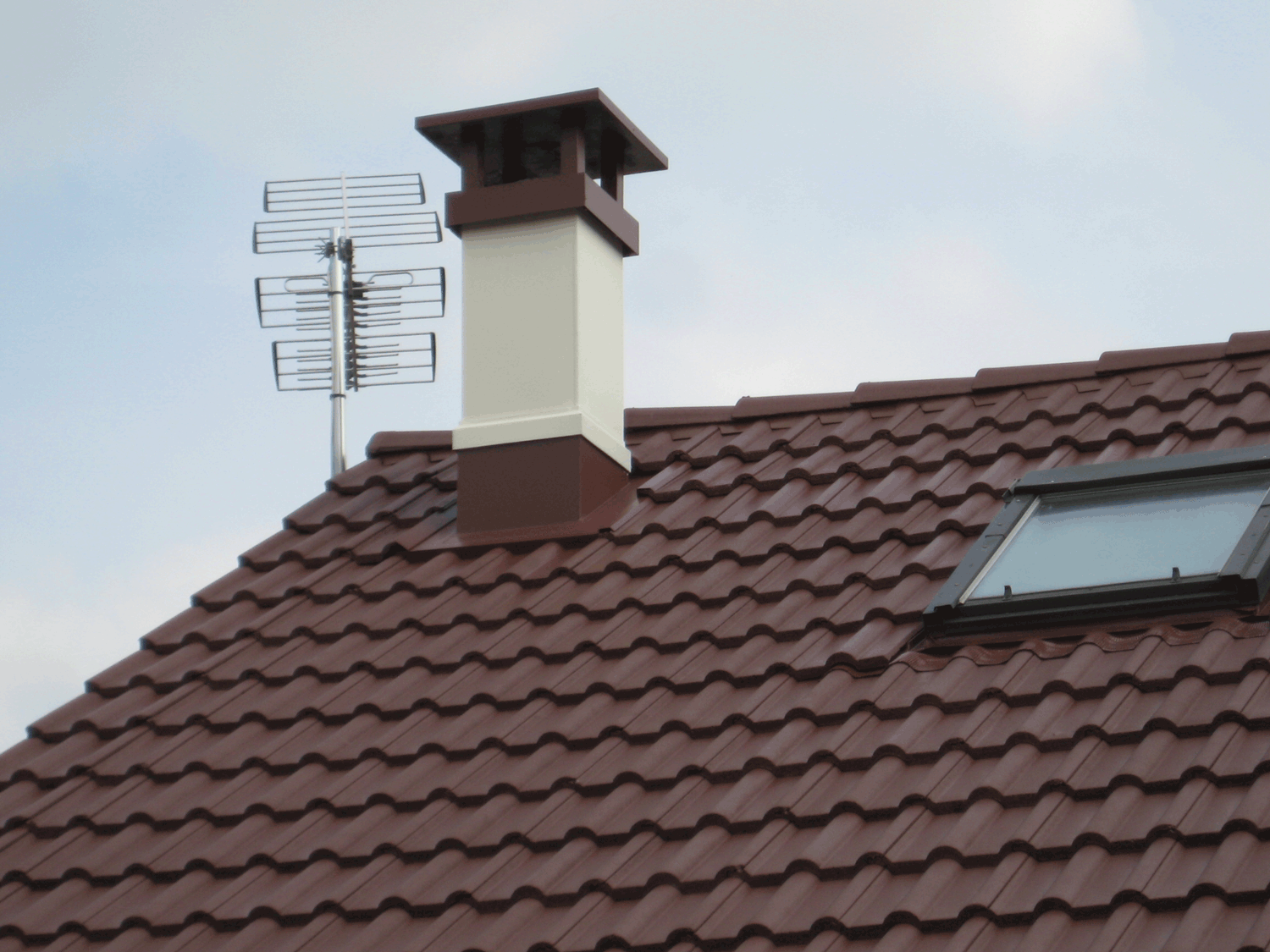 Toit en tuiles rouges, puits de lumière, cheminée et antenne de télévision sur fond de ciel nuageux.