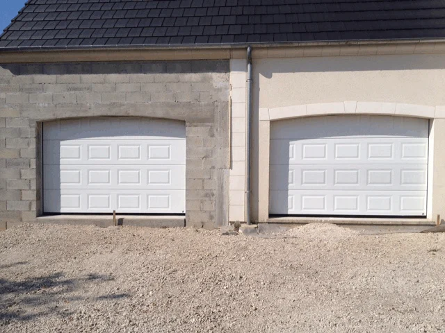 Deux portes de garage blanches encastrées dans une façade en pierre et en stuc, à côté d'une allée de gravier.