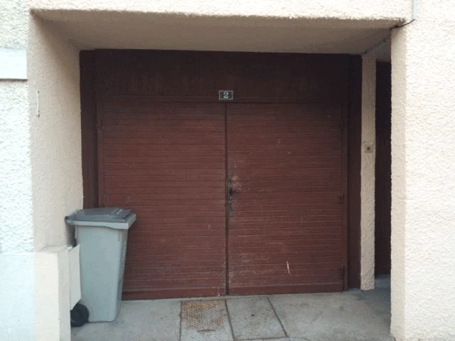 Portes de garage marron fermées à l'entrée d'un immeuble beige, avec une poubelle grise sur la gauche.