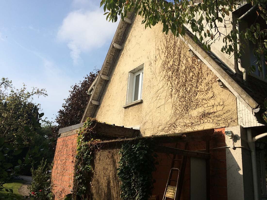 Une maison en stuc jaune avec un toit pentu et un porche recouvert de lierre dans un jardin ensoleillé