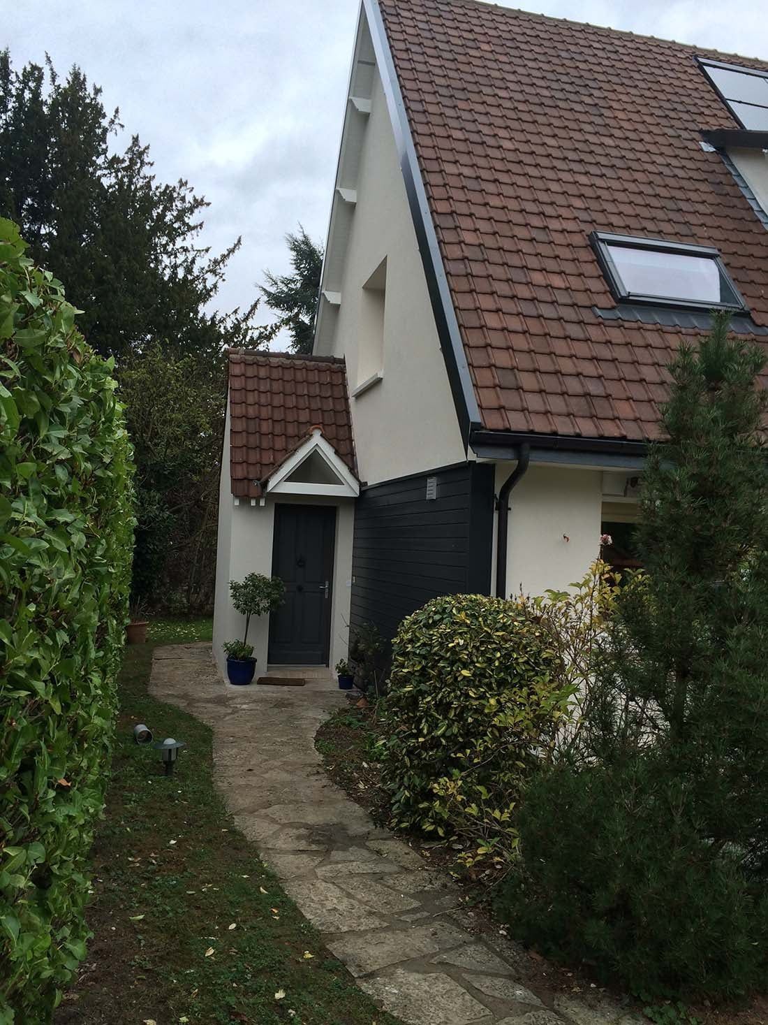 Un étroit chemin de jardin mène à l'entrée d'une maison aux murs blancs, à la porte noire et au toit de tuiles rouges.