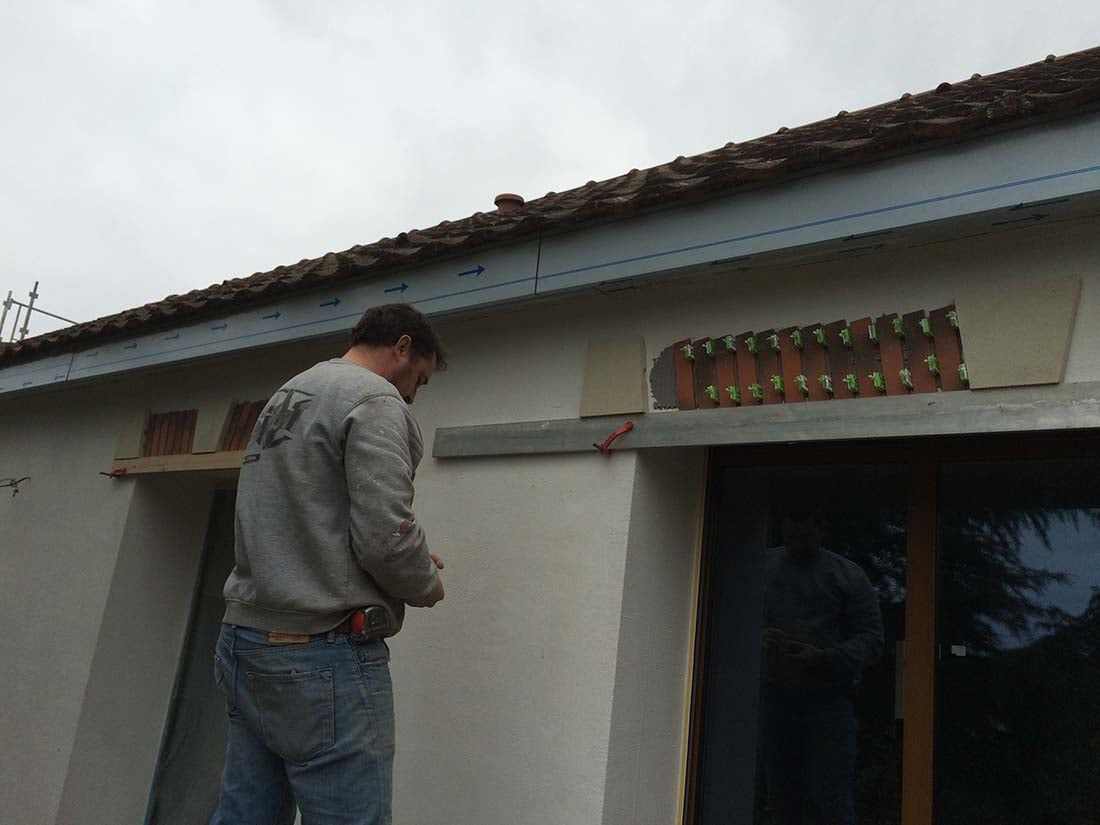 Un homme se tient debout près d'une maison blanche, travaillant près d'une porte sous un toit de tuiles.
