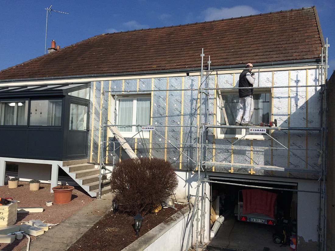 Extérieur de la maison avec échafaudages en cours de rénovation, toit de tuiles brunes et garage ouvert en dessous.
