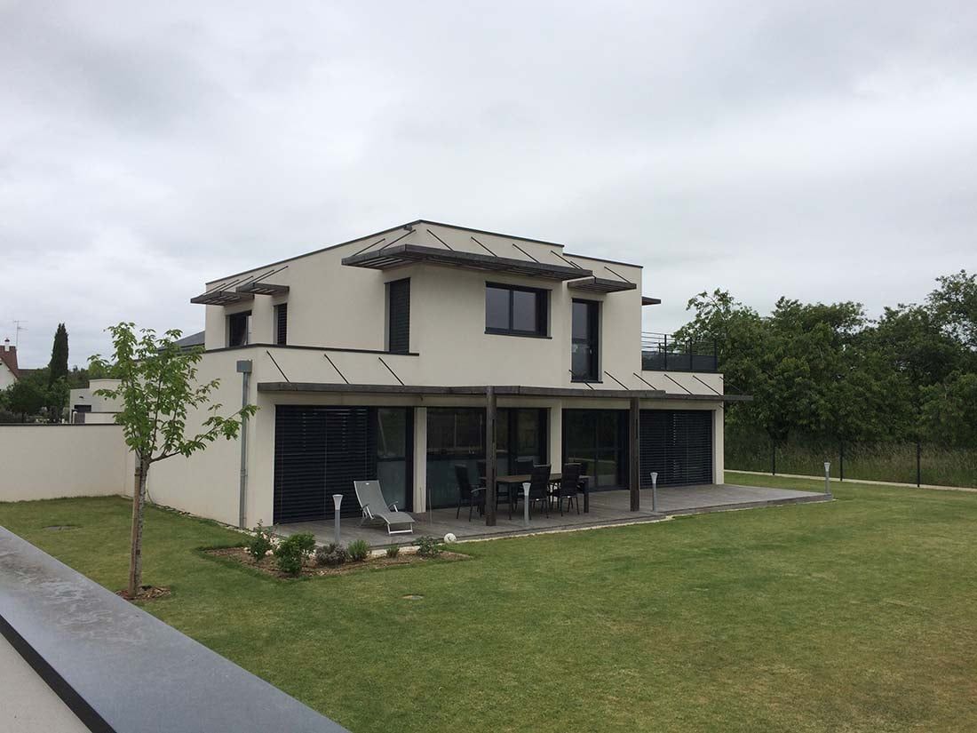 Maison beige à deux étages avec patio et grand jardin gazonné sous un ciel nuageux